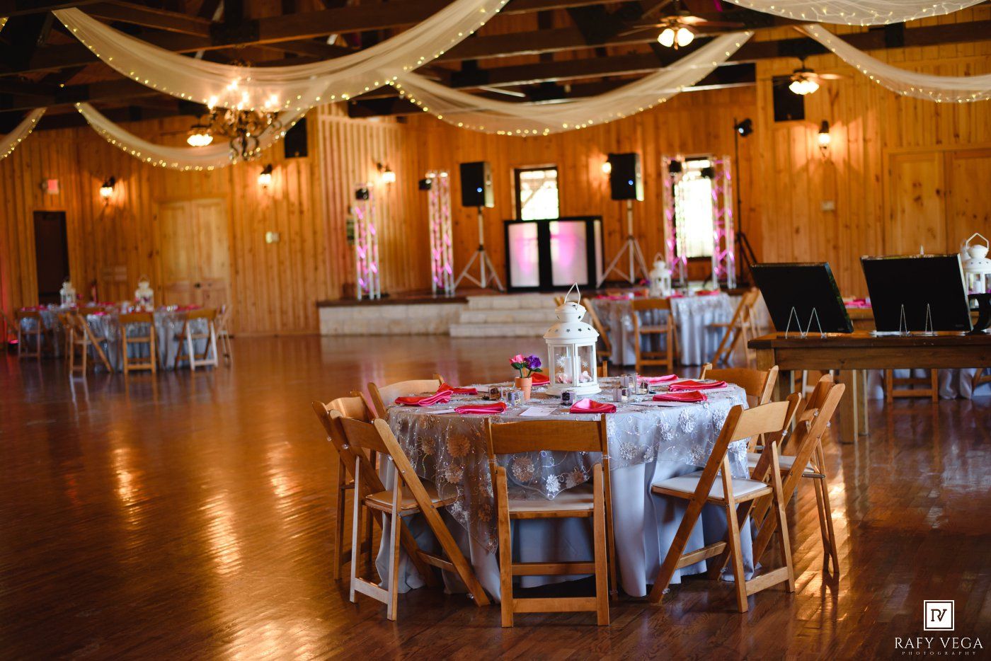 Rustic wedding reception venue with wooden walls, draped ceiling, round tables with white linens and red accents on hardwood floors.