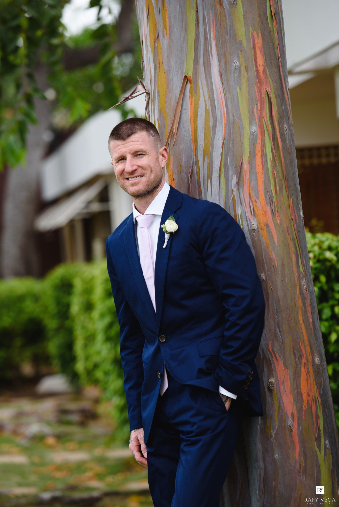 Groom in navy blue suit with boutonniere leaning against colorful rainbow eucalyptus tree outdoors.