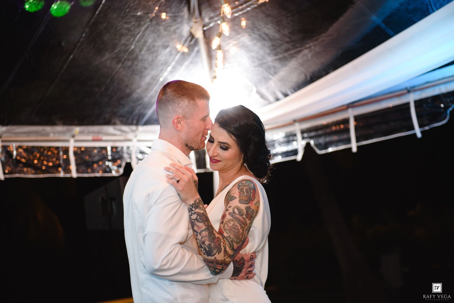 Tattooed bride and groom share first dance under glowing string lights at wedding reception.