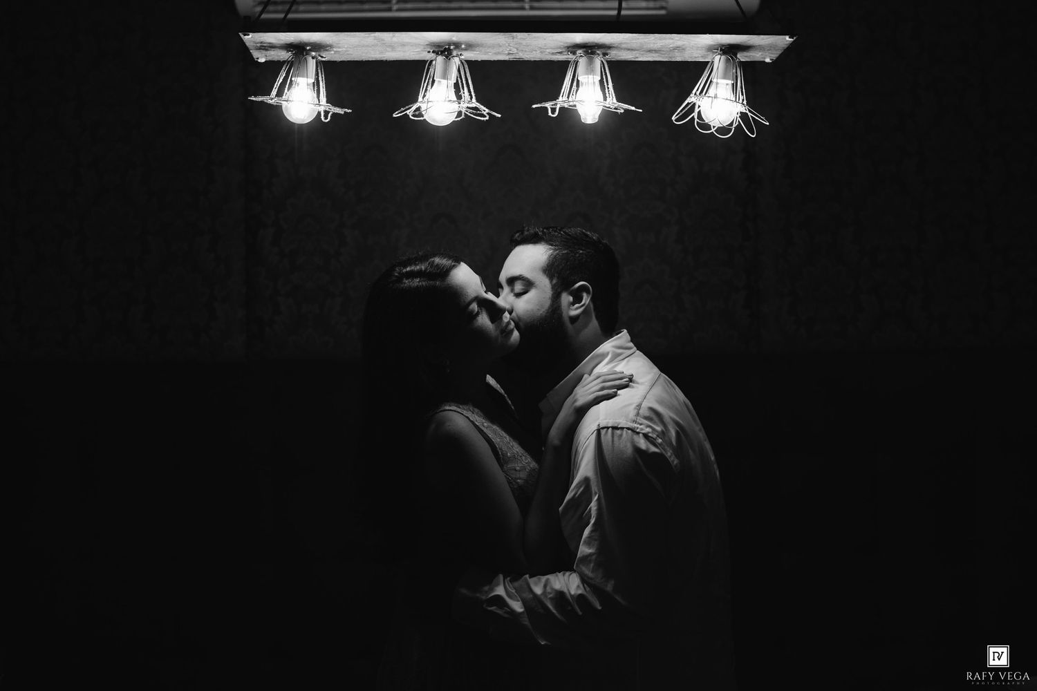 Black and white photo of a couple sharing a romantic kiss under vintage Edison bulb lights in a dark setting.