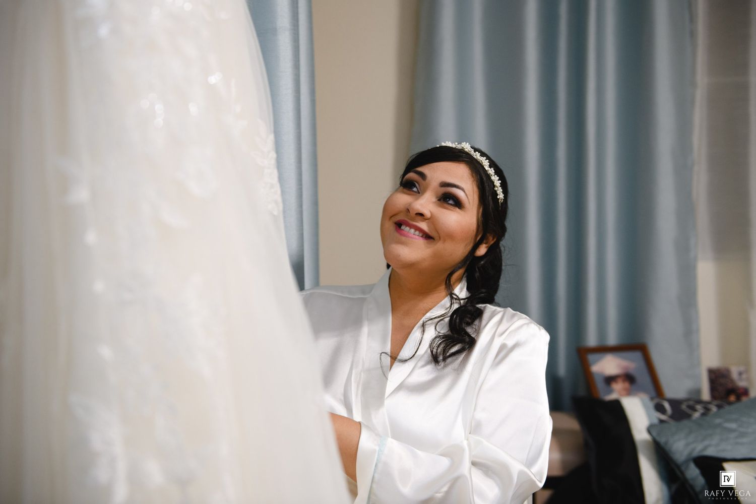 Bride in white satin robe smiling up at her wedding gown hanging in a softly lit bridal suite.