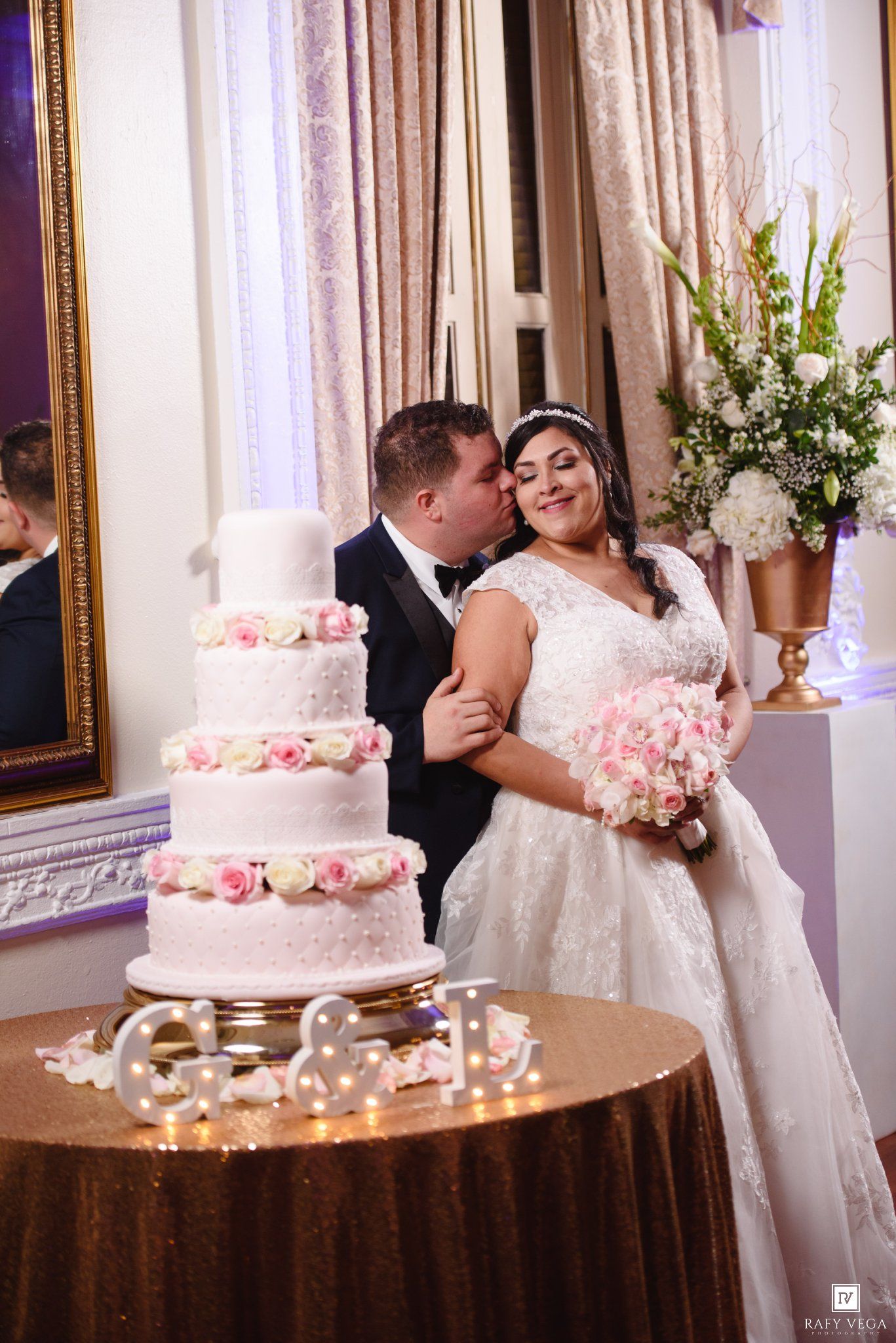 Bride and groom share a kiss beside a four-tier white wedding cake adorned with pink roses at their elegant reception.
