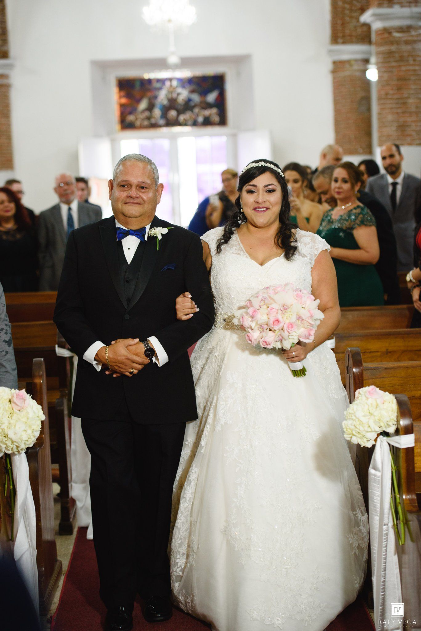Bride in white gown walks down church aisle with escort, holding pink rose bouquet during wedding ceremony.