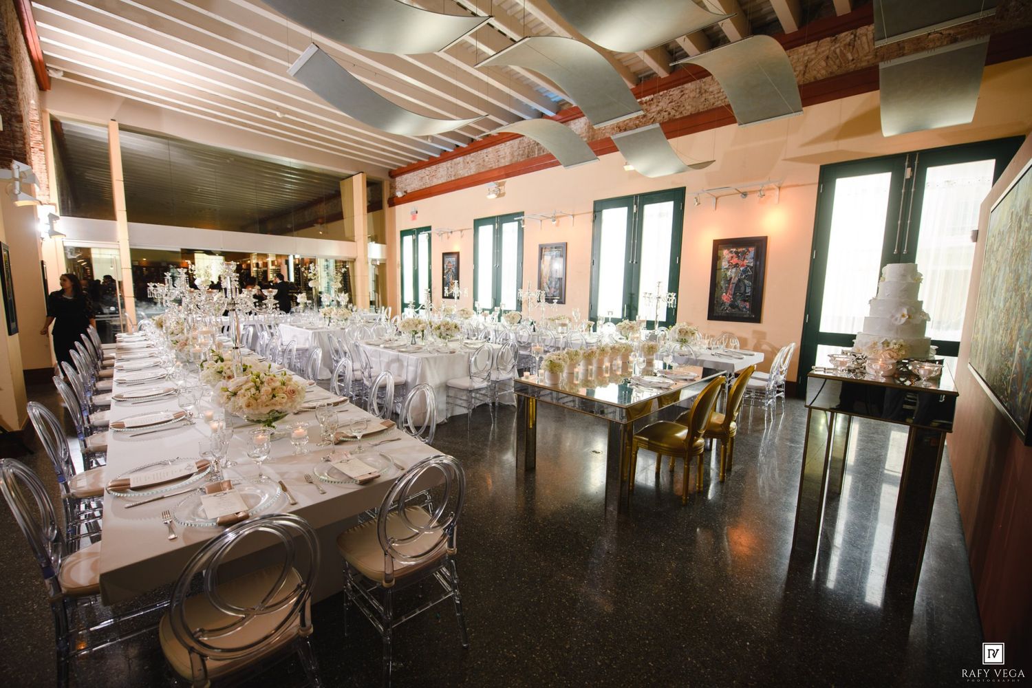 Elegant wedding reception hall with long white-draped tables, ghost chairs, and a tiered wedding cake on display.