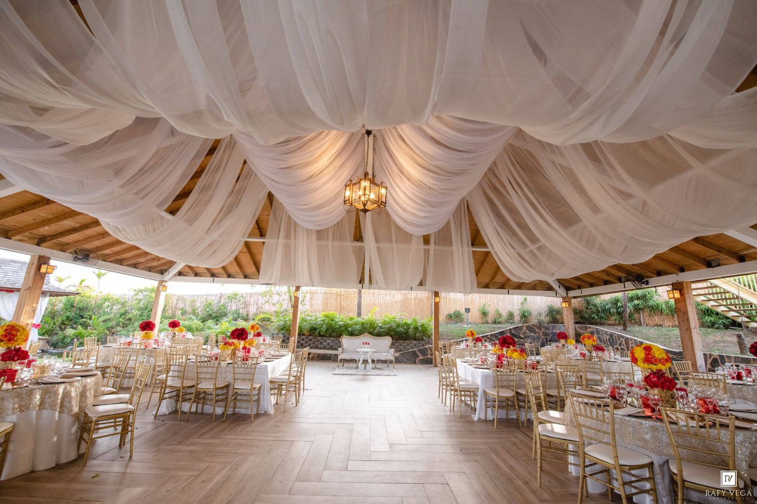 Elegant outdoor wedding reception venue with white draped ceiling, chandelier, gold chiavari chairs, and vibrant floral centerpieces.