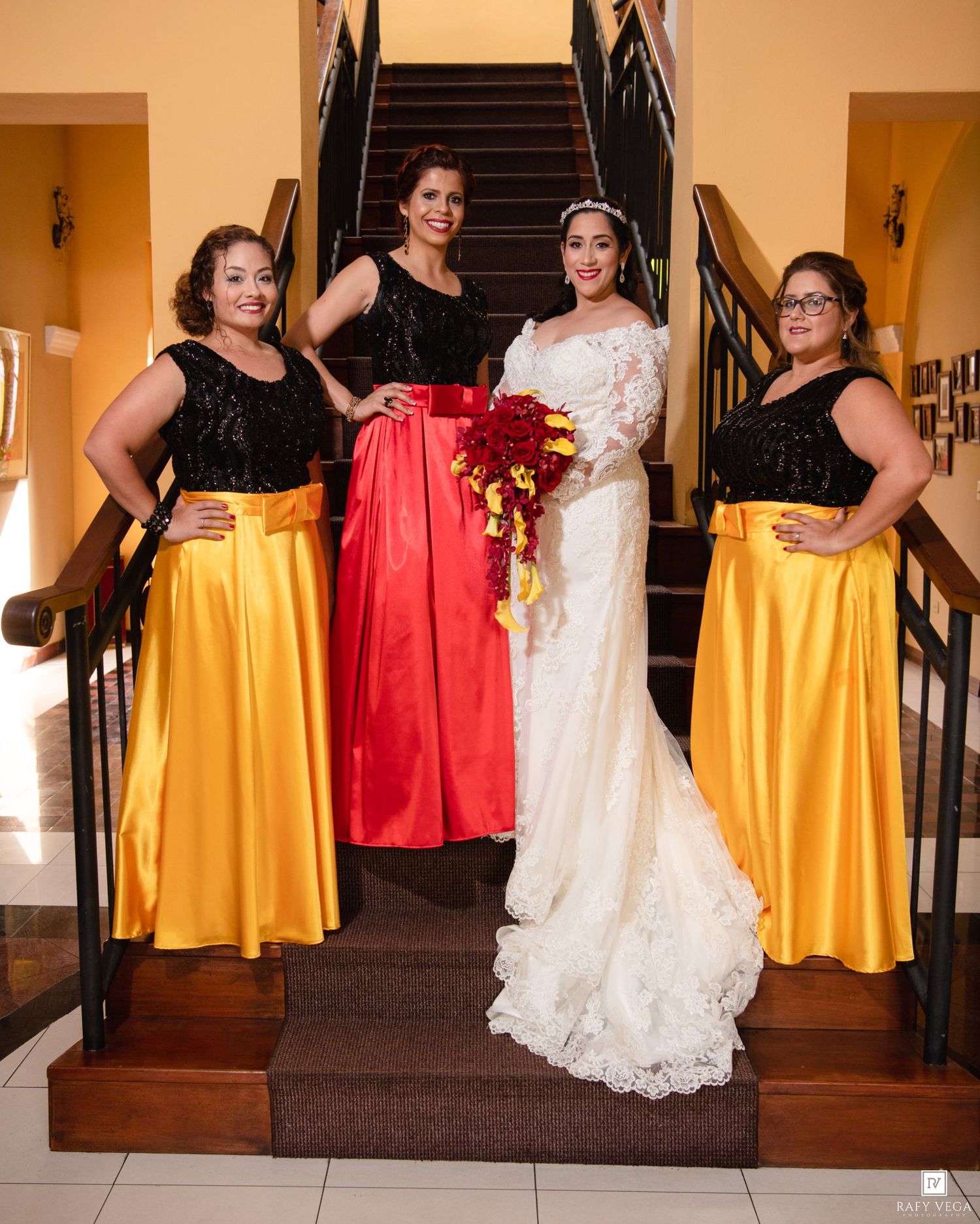Bride in white lace gown with three bridesmaids in black tops and colorful skirts posing on staircase.