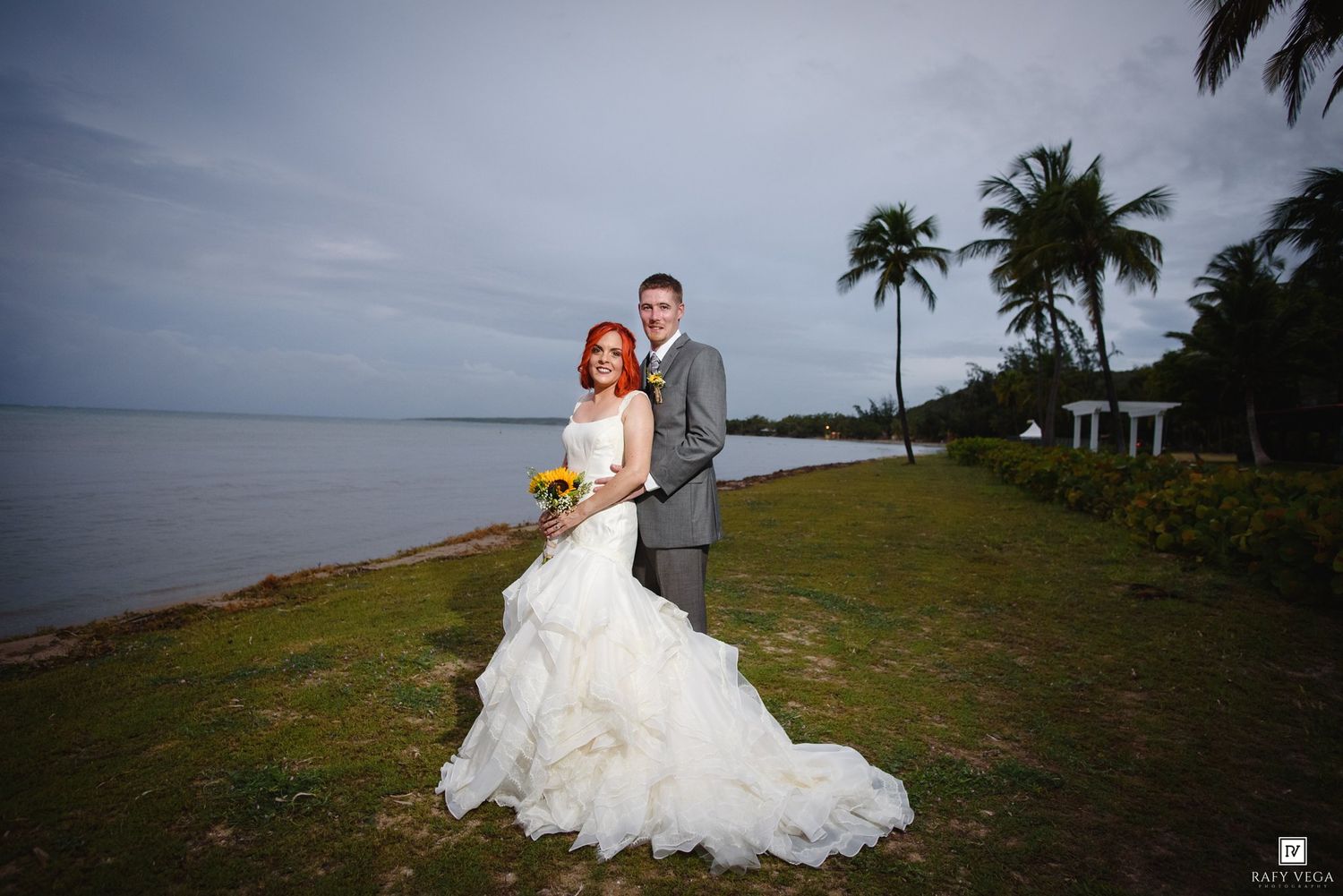 Bride and groom pose together on a lush waterfront lawn with palm trees and ocean views on their wedding day.