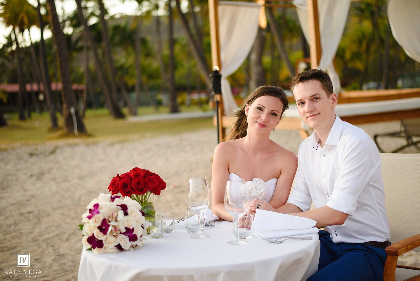 Bride and groom sitting together at a decorated beach table with red roses bouquet during outdoor wedding ceremony.