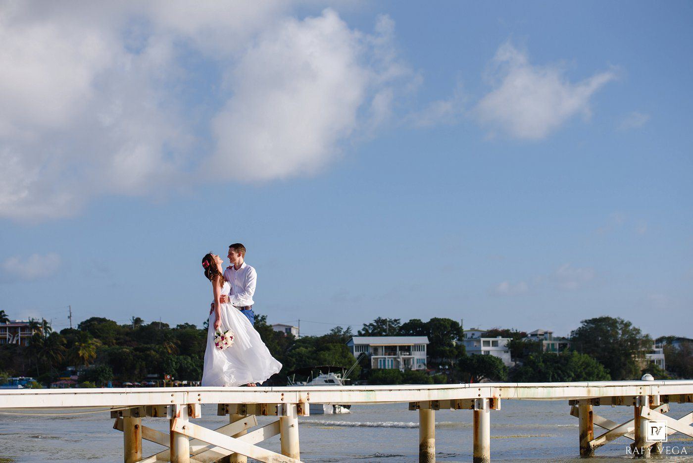 A bride and groom share a romantic kiss on a white pier overlooking calm blue waters under a partly cloudy sky.