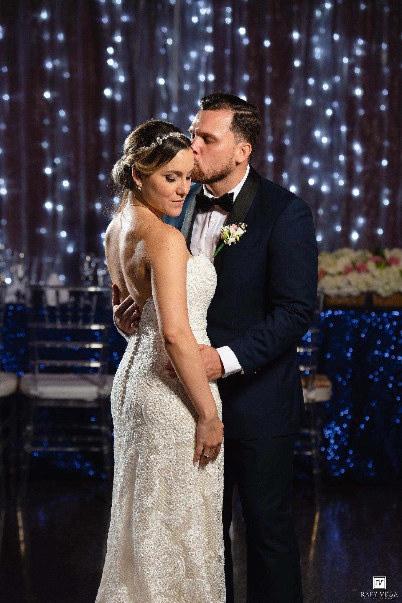 Groom in tuxedo kissing bride in lace gown on forehead against sparkling fairy light backdrop at wedding.