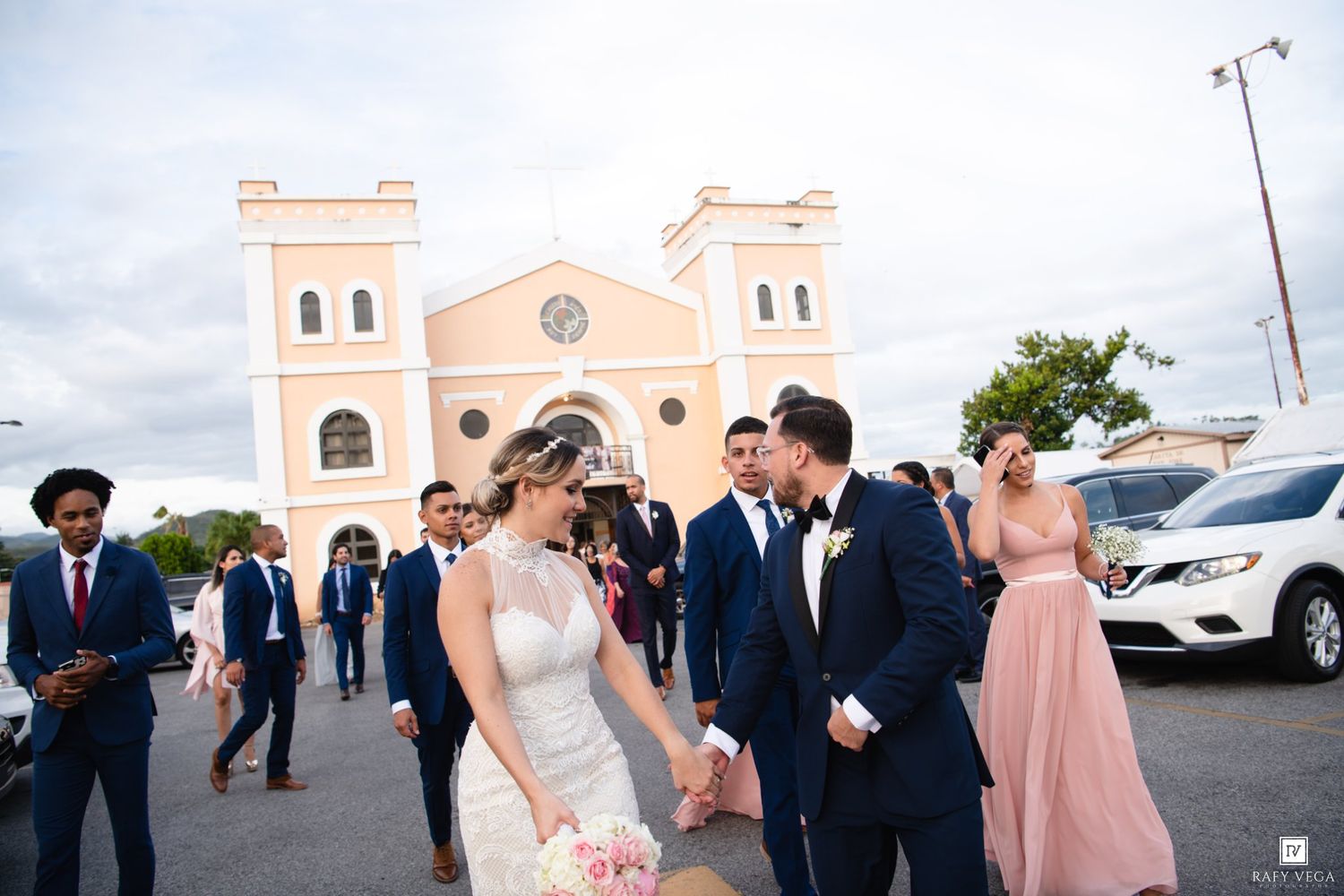 Bride and groom share a romantic moment outside a pink church surrounded by wedding party guests in formal attire.