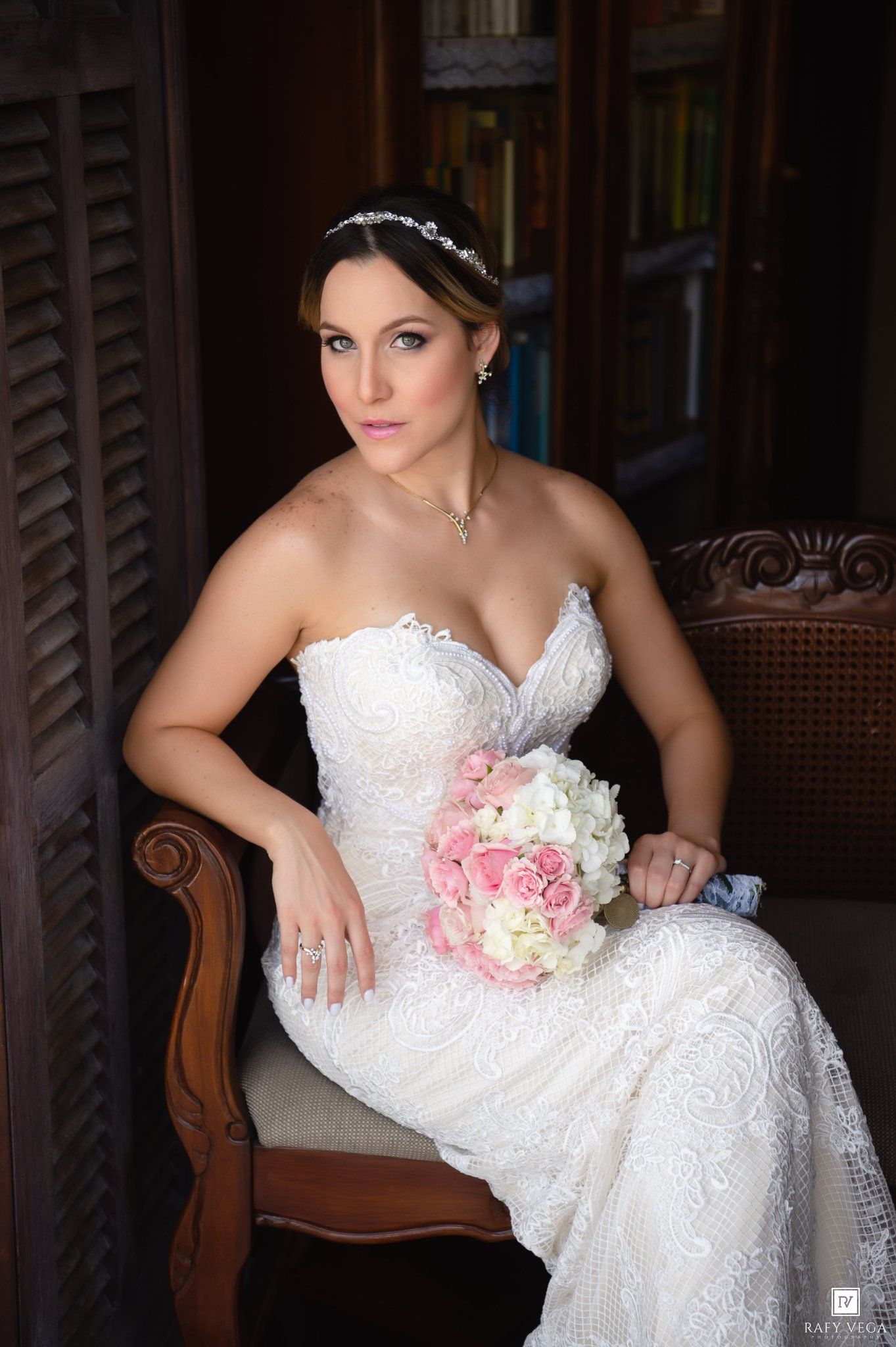 Elegant bride in strapless lace wedding gown seated in wooden chair holding pink and white rose bouquet with crystal headpiece.