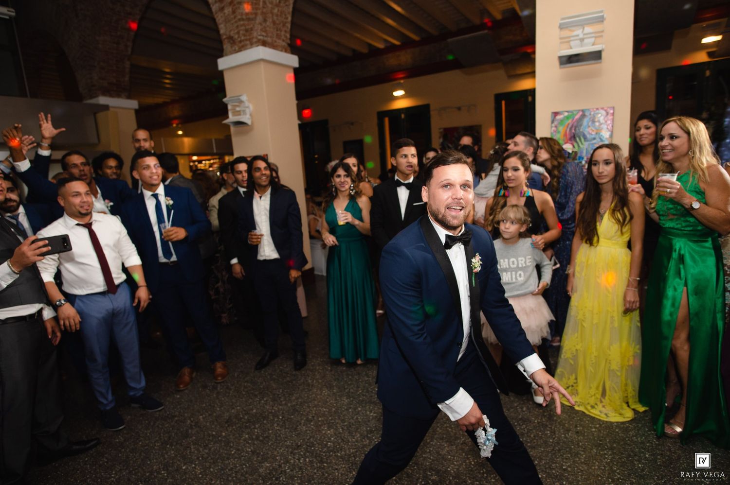 Groom in navy tuxedo dancing at wedding reception surrounded by elegantly dressed guests in formal attire.