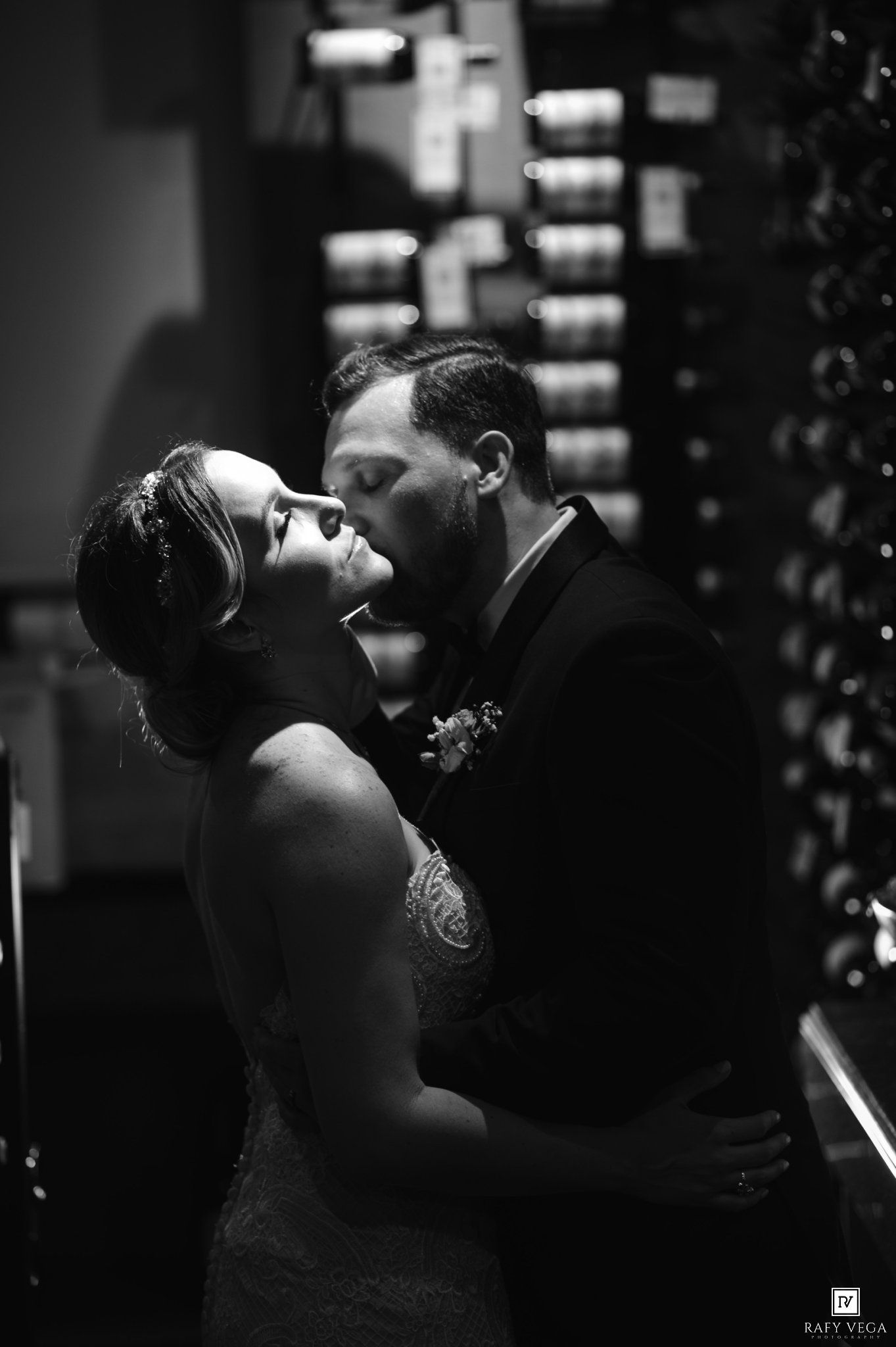 Black and white wedding photo of bride and groom sharing a romantic kiss near a wine rack.