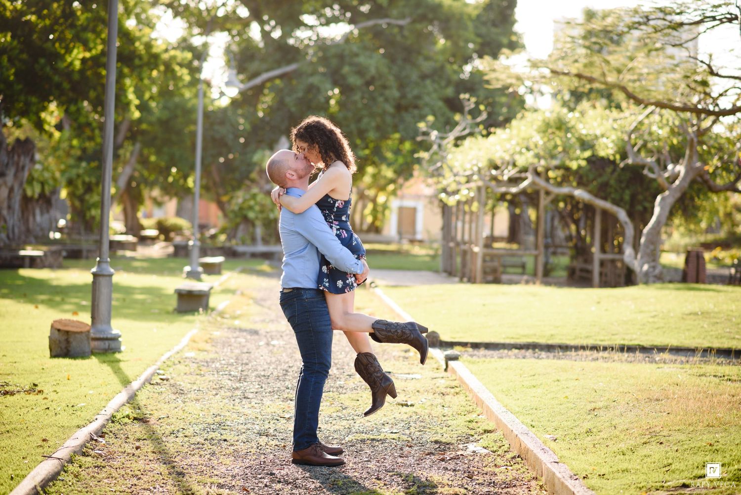 Couple embracing on railway tracks in a sunny park, man lifting woman in cowboy boots during romantic outdoor photoshoot.