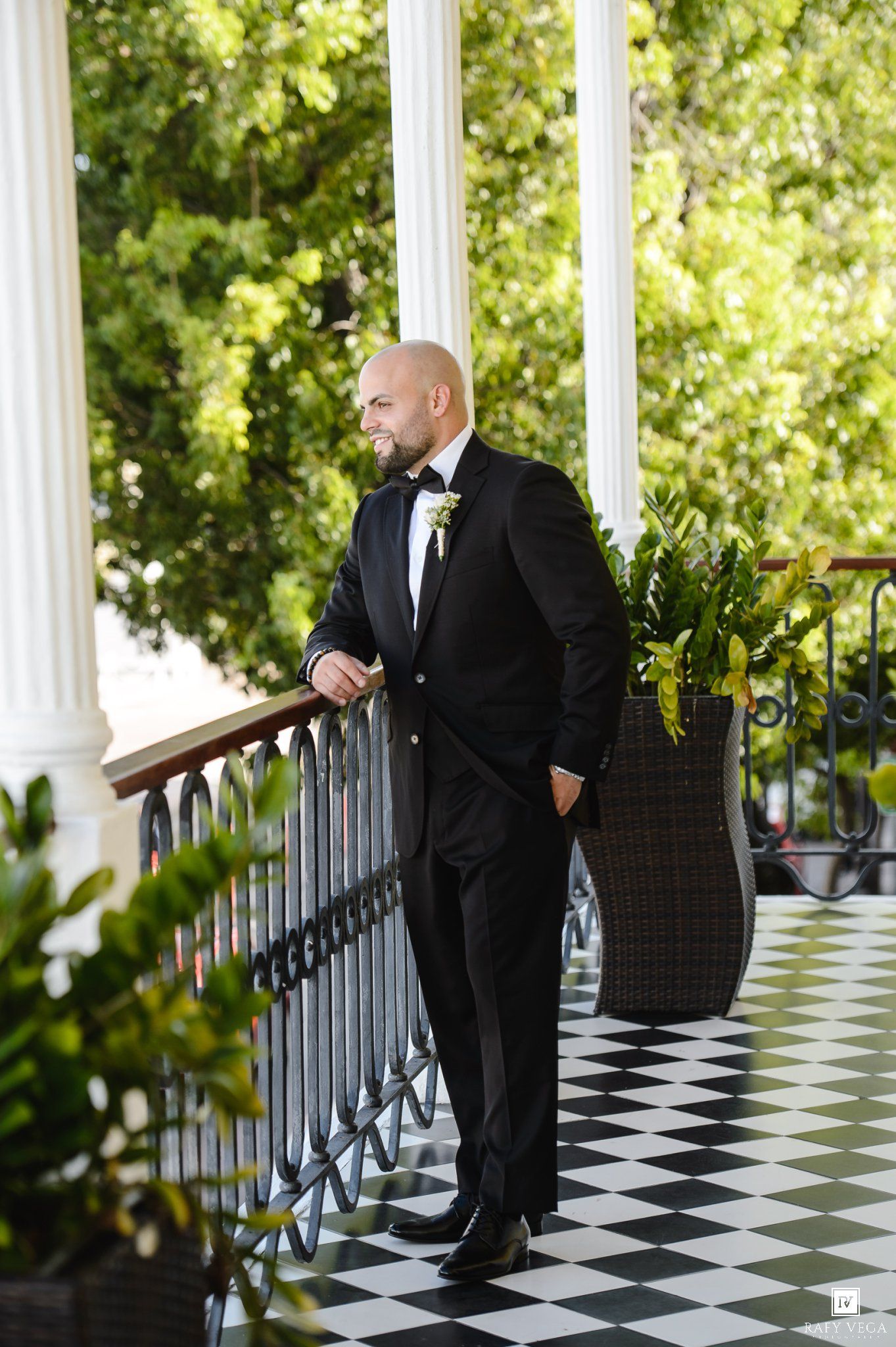 Groom in black tuxedo with boutonniere stands on checkered porch with white columns and lush greenery.