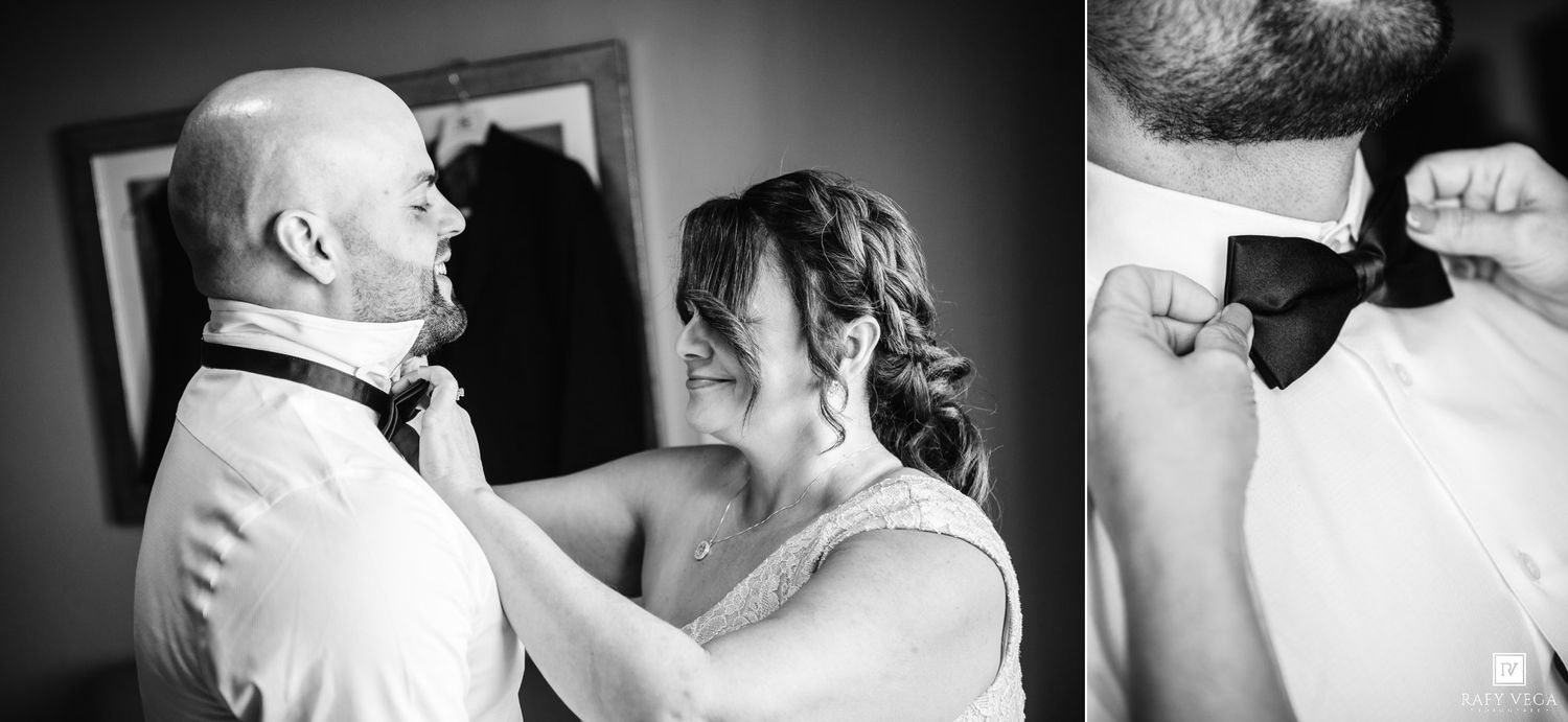 Black and white photo of a bride adjusting the groom's bow tie while smiling on their wedding day.
