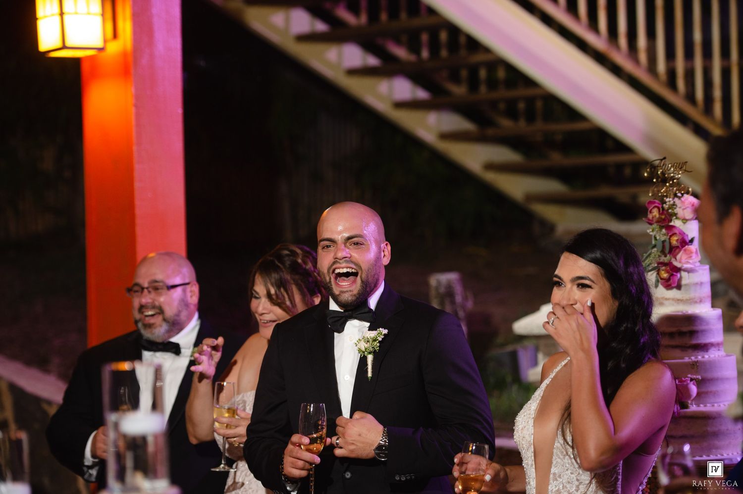 Groom laughing joyfully with bride and guests at elegant wedding reception near staircase with wedding cake.