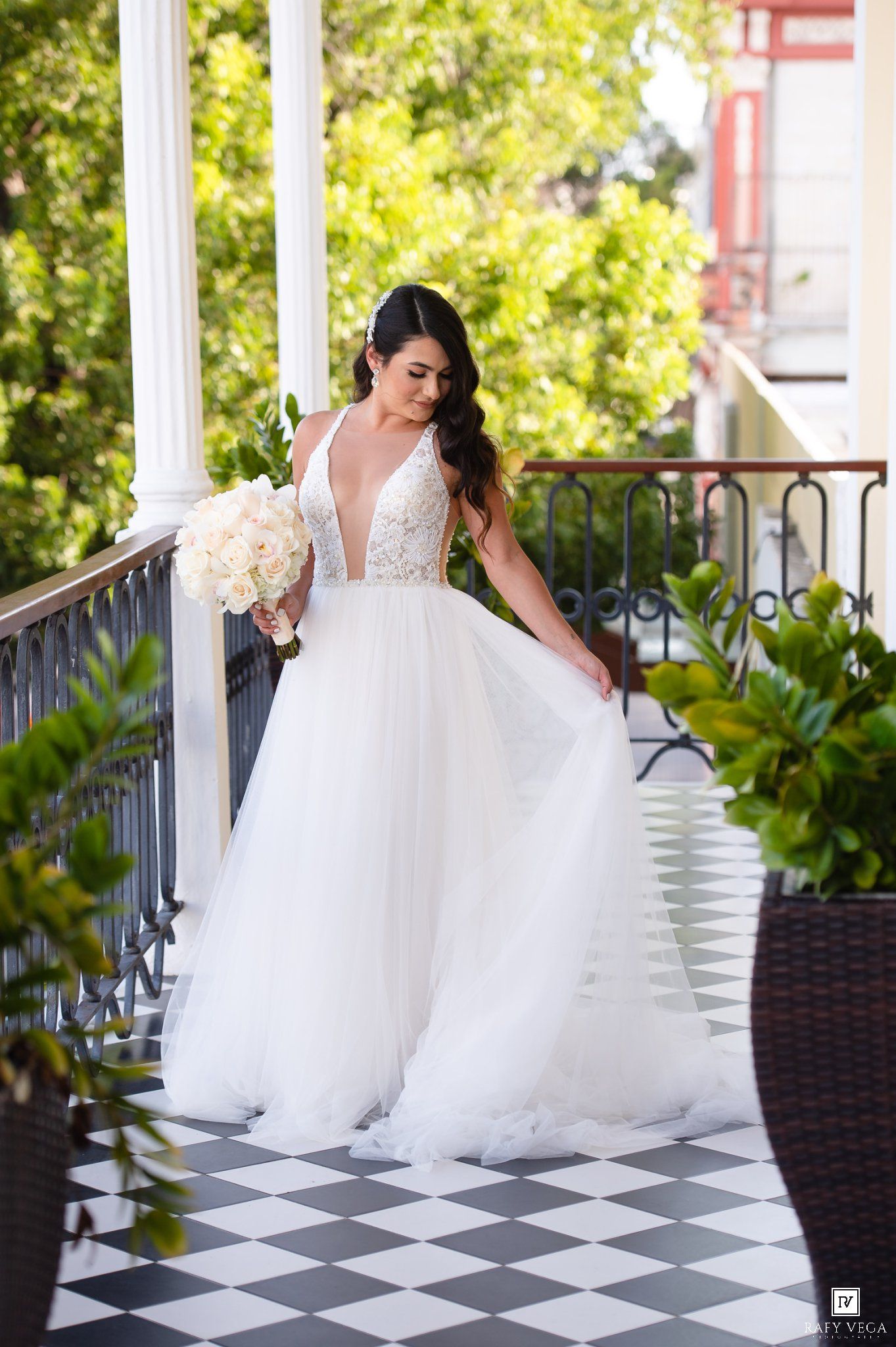 Bride in a deep V-neck tulle ball gown holding white bouquet on a elegant balcony with checkered floor.
