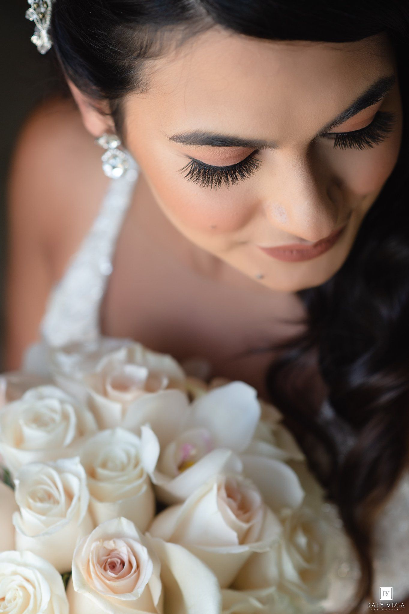 Bride with dramatic lashes and pearl earrings gazes down at a bouquet of white roses on her wedding day.