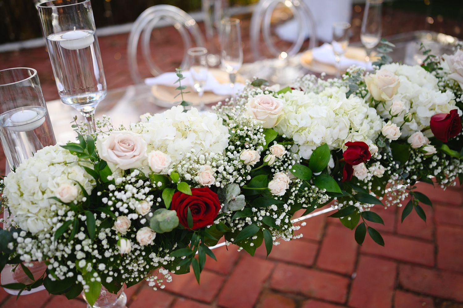 Elegant wedding table centerpiece with white hydrangeas, red and blush roses, and baby's breath on a brick patio.