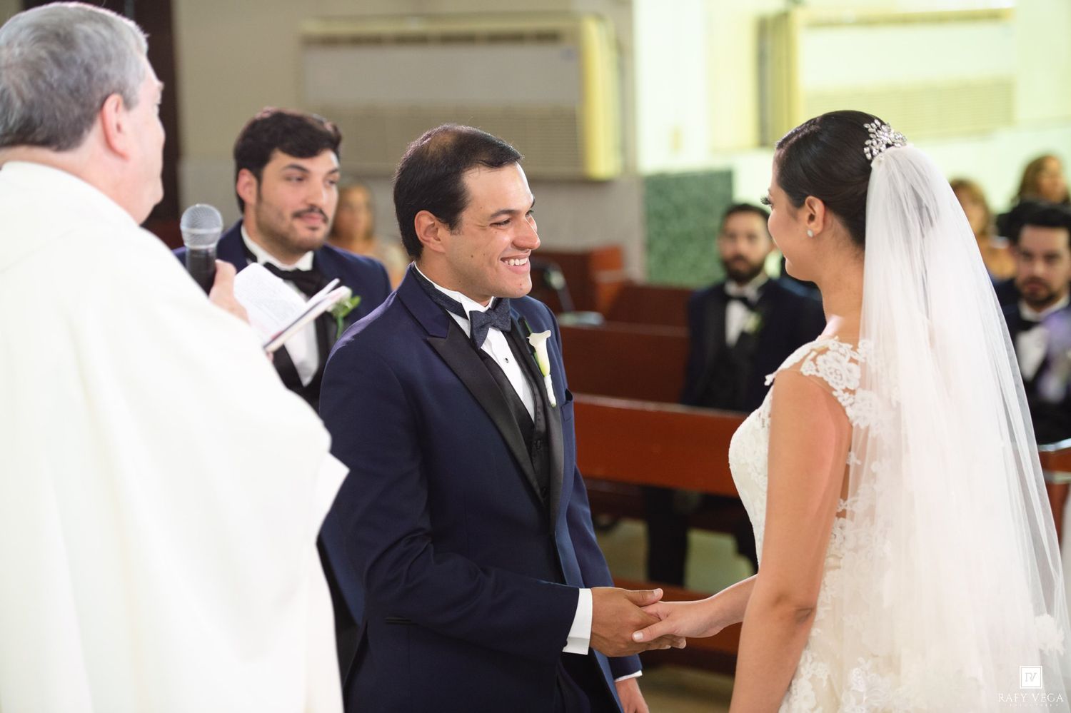 Smiling groom holds brides hands at altar during elegant church wedding ceremony with officiant.