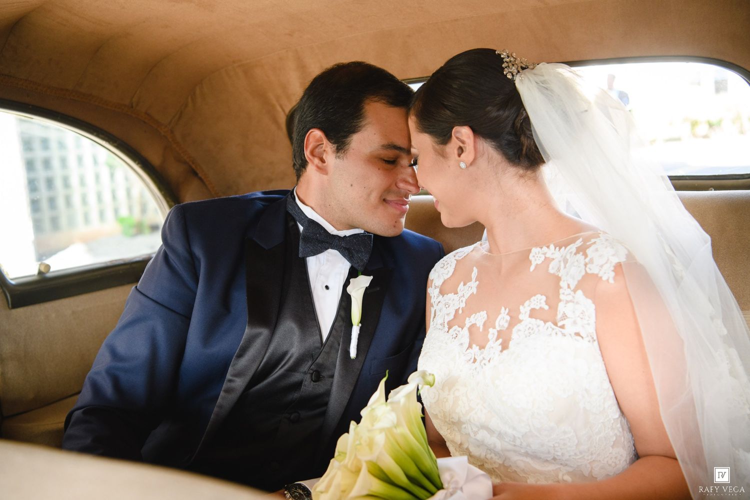 Bride and groom share a romantic moment in a vintage car, bride in lace gown with veil holding white bouquet.