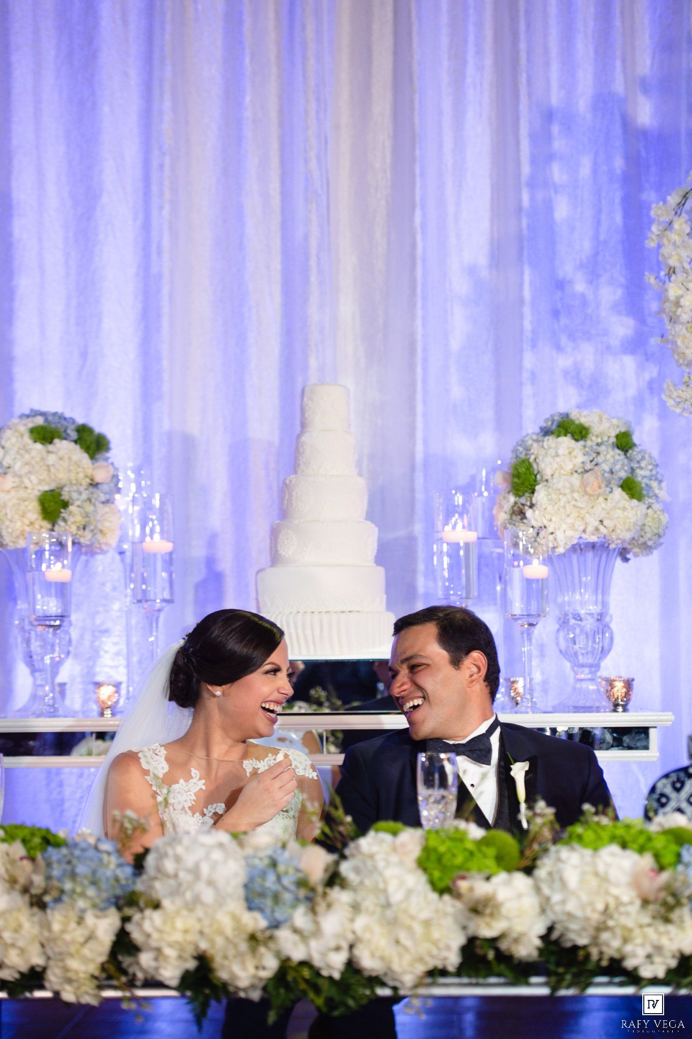 Bride and groom laughing at sweetheart table with white floral arrangements and tiered wedding cake backdrop.