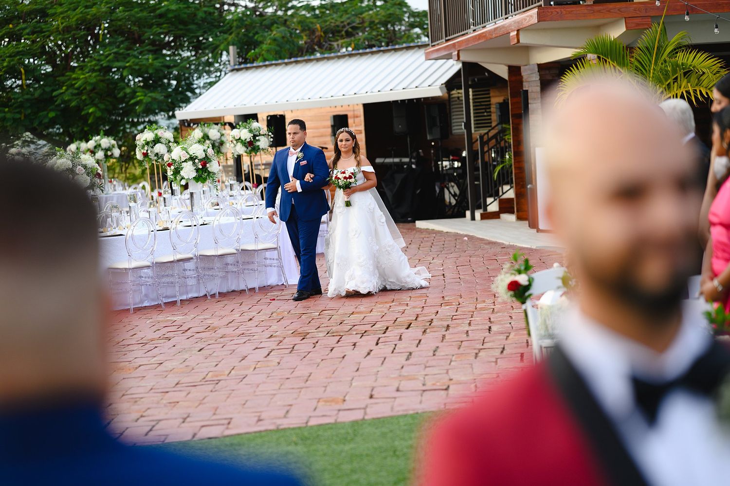 Bride and groom walking together at outdoor wedding reception with white floral decorations and brick patio venue.
