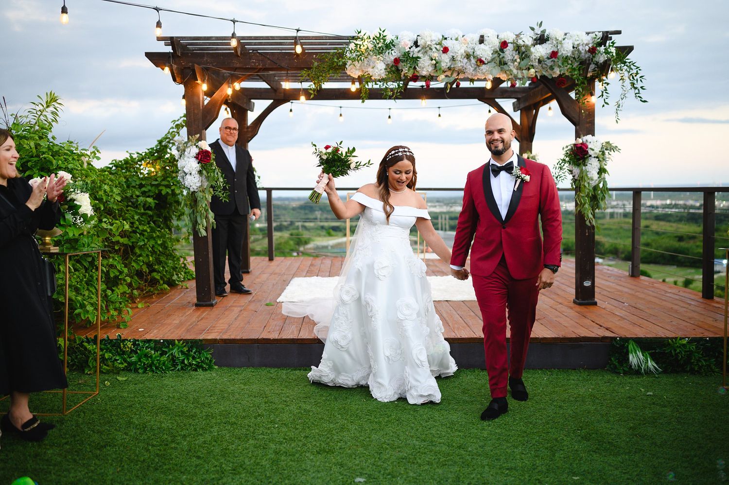 Bride and groom celebrate after outdoor rooftop wedding ceremony under floral pergola with string lights.