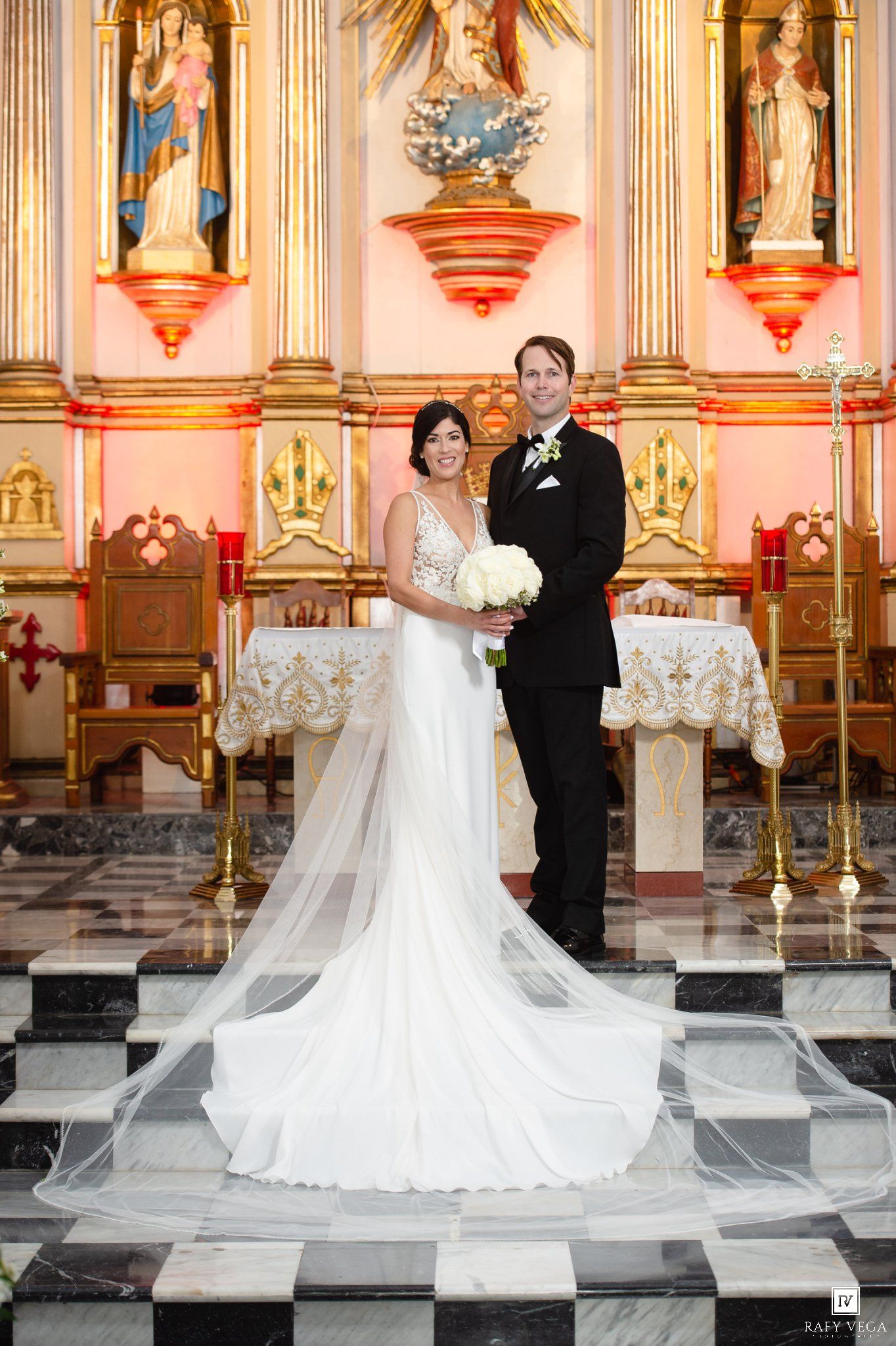 Bride and groom pose at church altar on checkered floor, bride in white gown with veil, groom in black tuxedo.