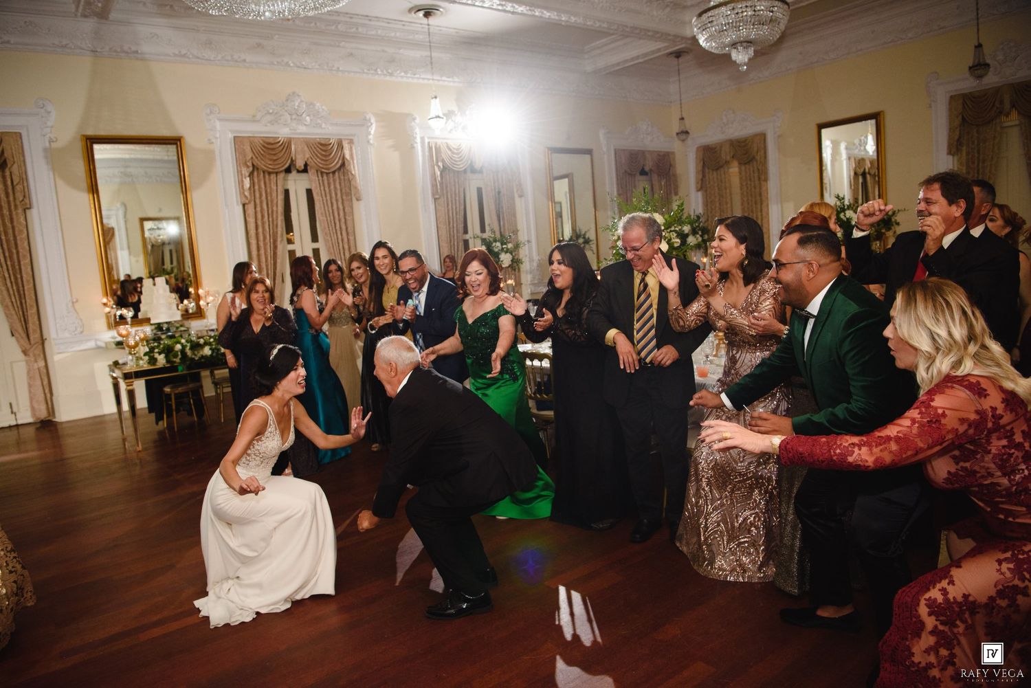 Elegant wedding reception in a grand ballroom with guests watching a couple during a lively garter or bouquet toss tradition.
