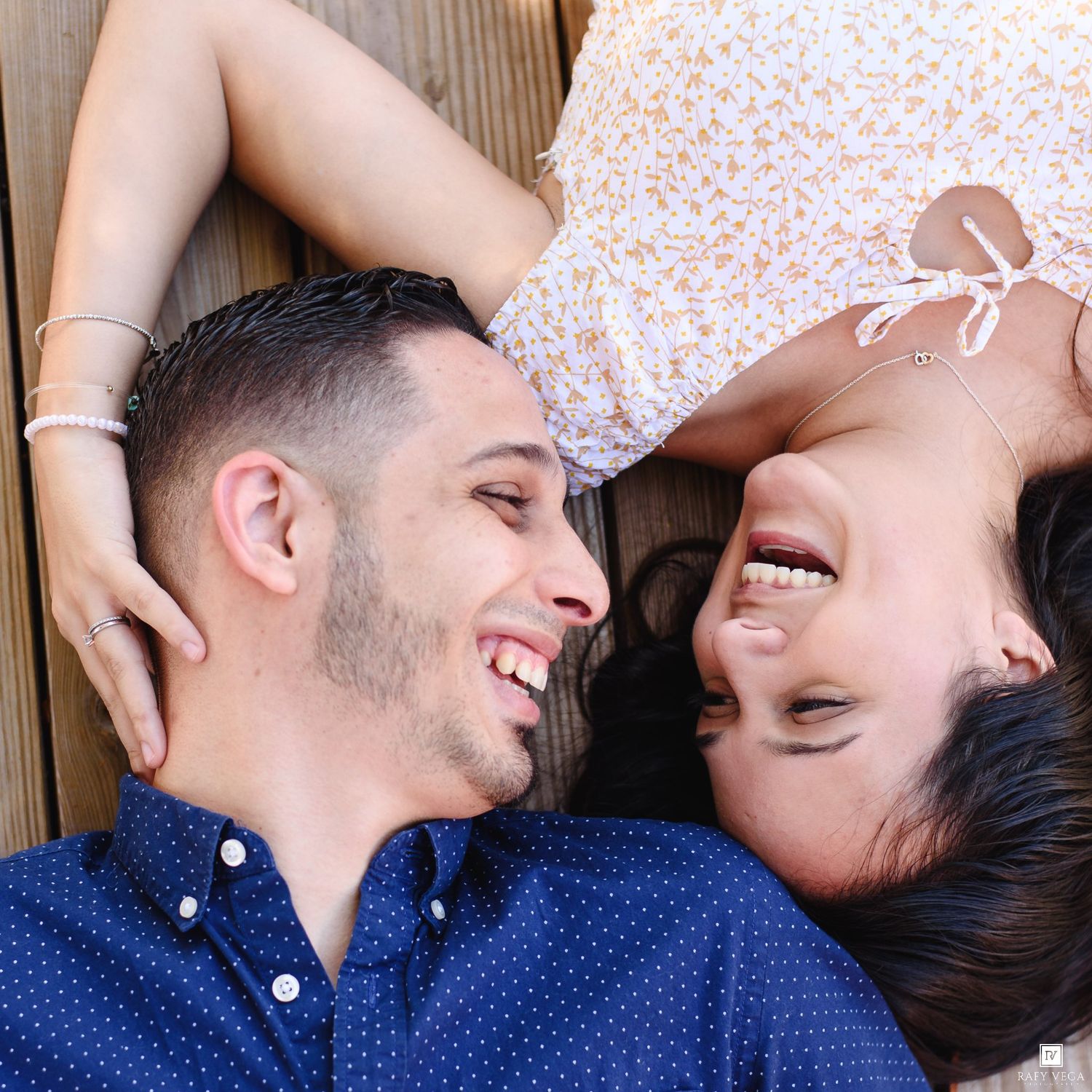 A smiling couple lying on a wooden deck, gazing at each other, the man in a blue polka-dot shirt and woman in a floral dress.