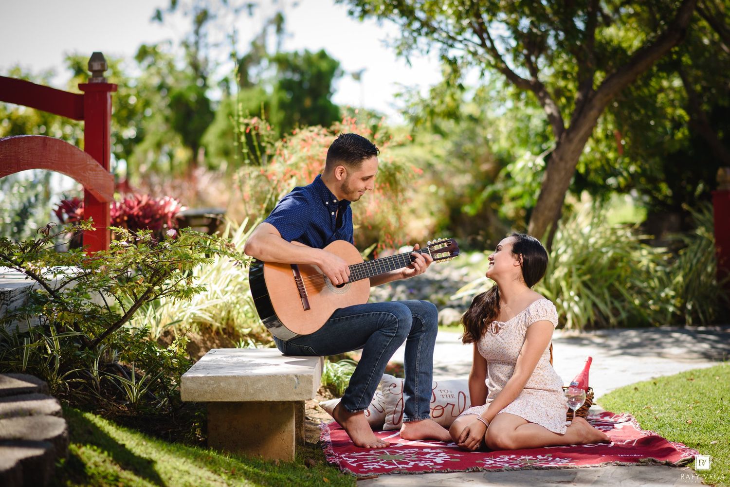 Man playing acoustic guitar for woman on picnic blanket in lush garden park setting.
