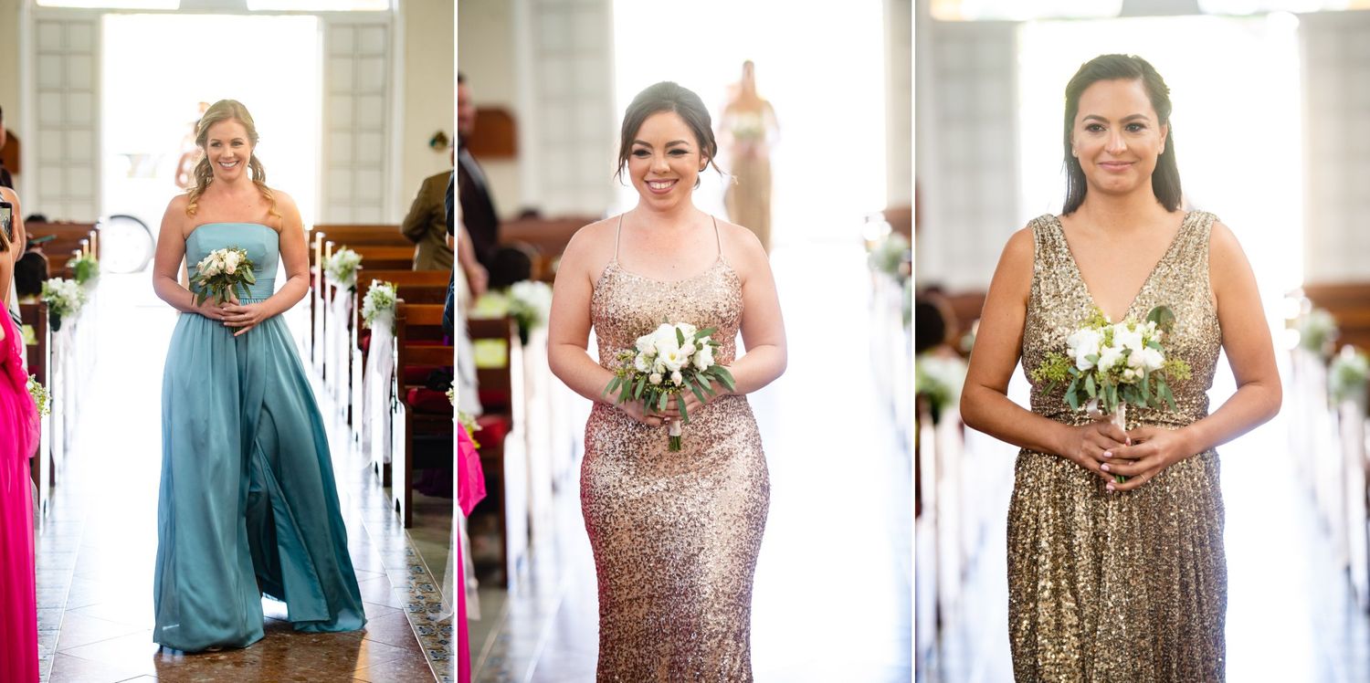 Three bridesmaids in elegant gowns walking down the aisle, wearing blue, rose gold, and gold sequin dresses.