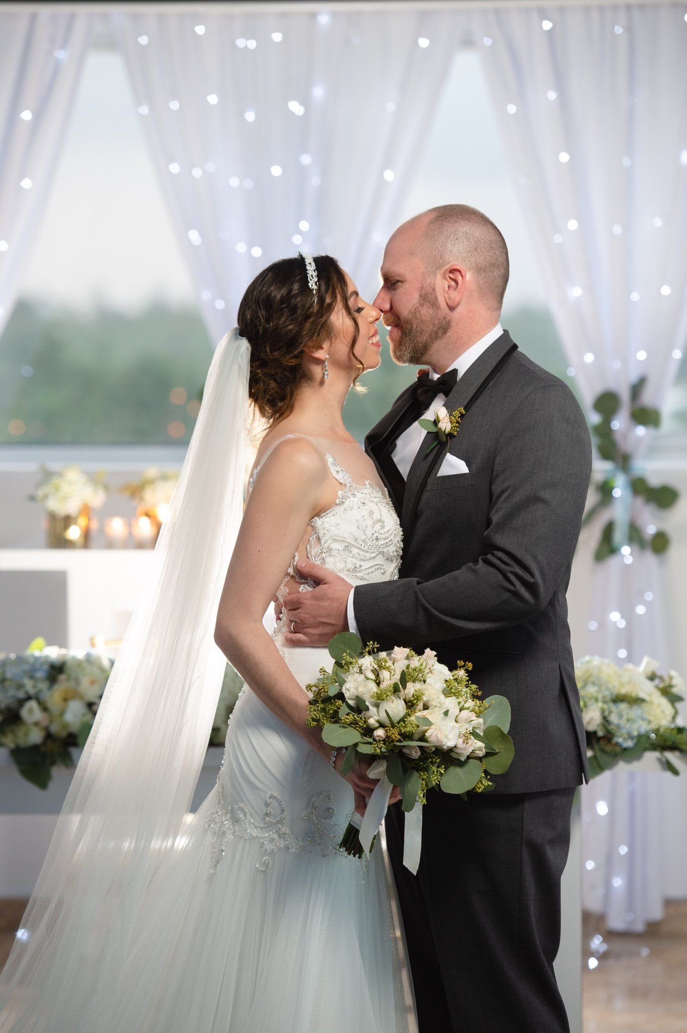 Bride and groom share a romantic kiss at the altar, surrounded by fairy lights and white floral arrangements.