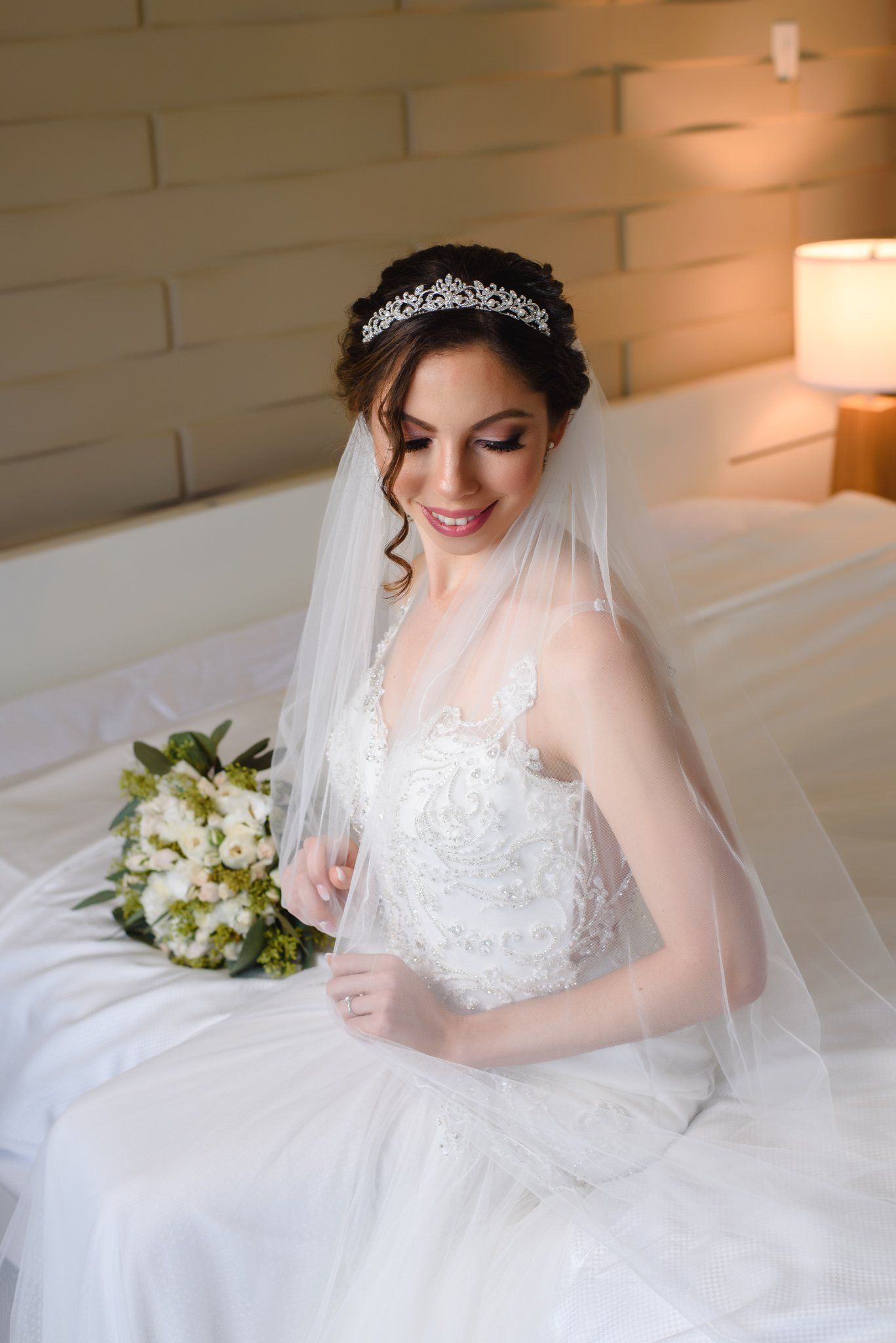 Smiling bride in lace wedding dress and tiara sits on bed holding white floral bouquet with veil draped over shoulders.