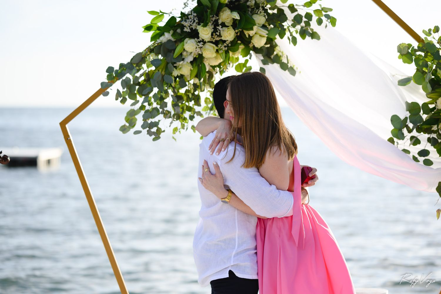 Couple embracing under a floral arch by the sea at a romantic outdoor wedding ceremony.