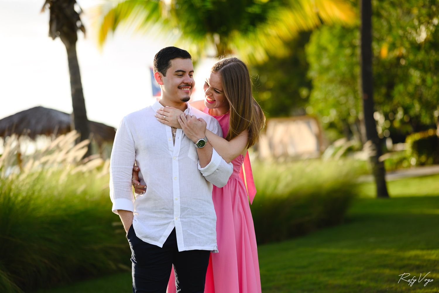 Couple embracing outdoors in tropical garden, woman in pink dress hugging man in white shirt at sunset.