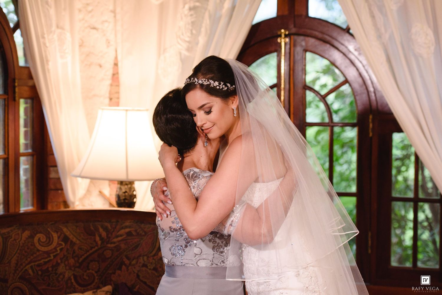 Bride in white gown and veil shares emotional hug with bridesmaid in silver dress.