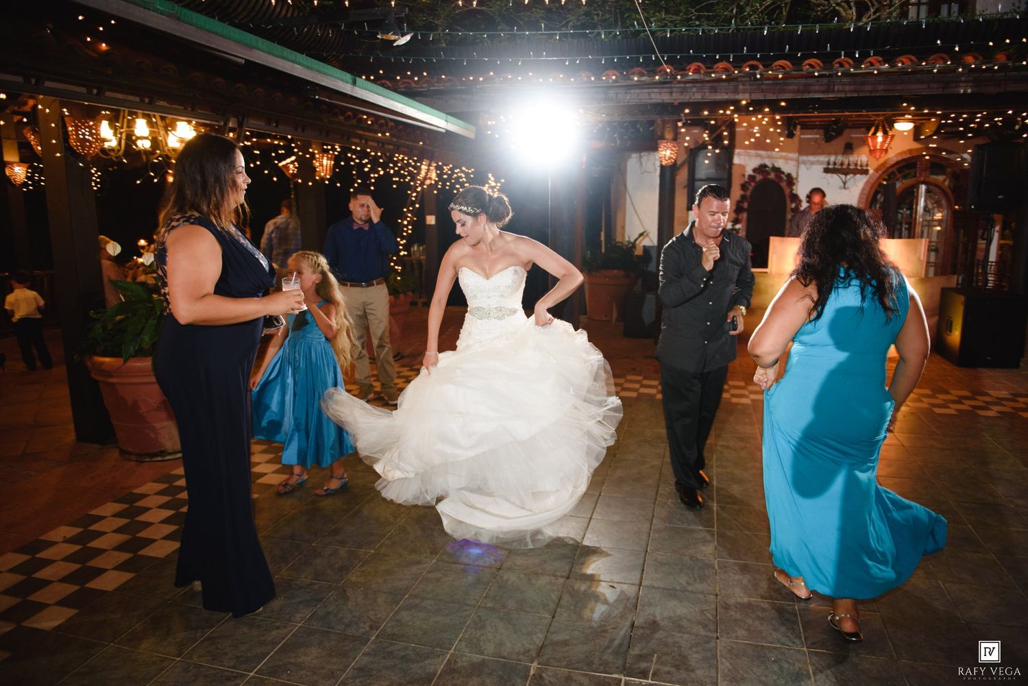 Bride in white gown dances joyfully with guests in teal dresses at an elegant indoor wedding reception.