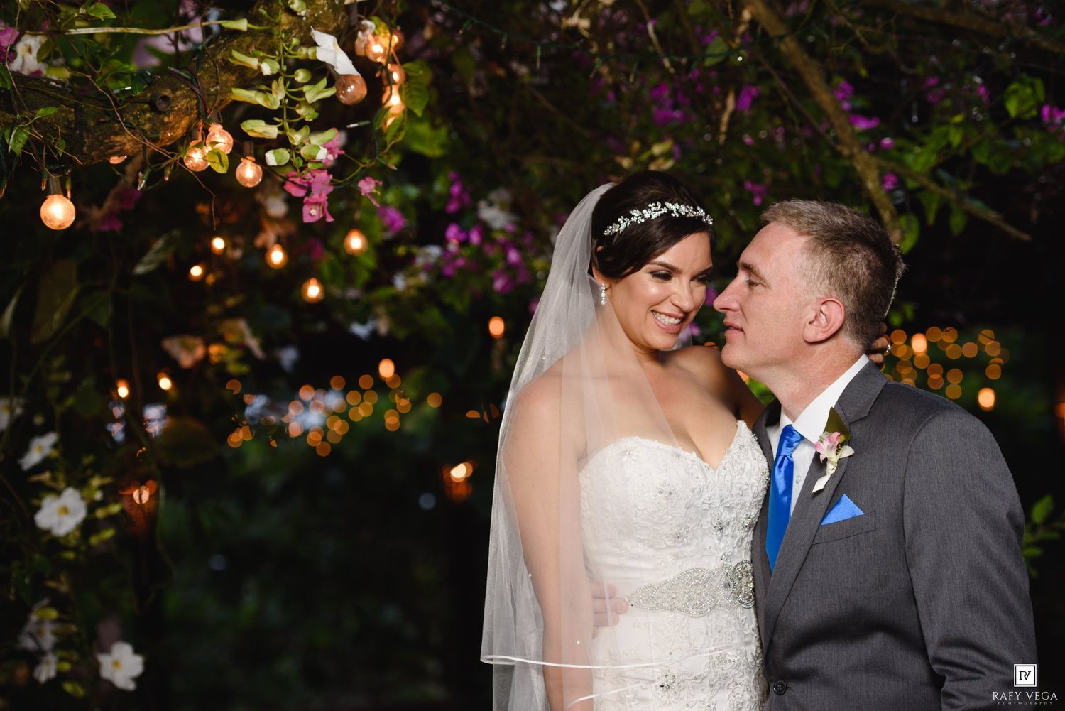 Bride and groom share a romantic moment under floral arch with twinkling lights at outdoor wedding.