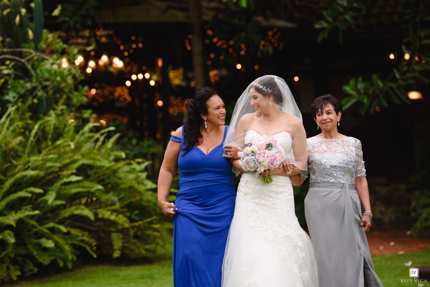 Bride in white gown and veil walks with two women in blue and gray dresses at an outdoor wedding.