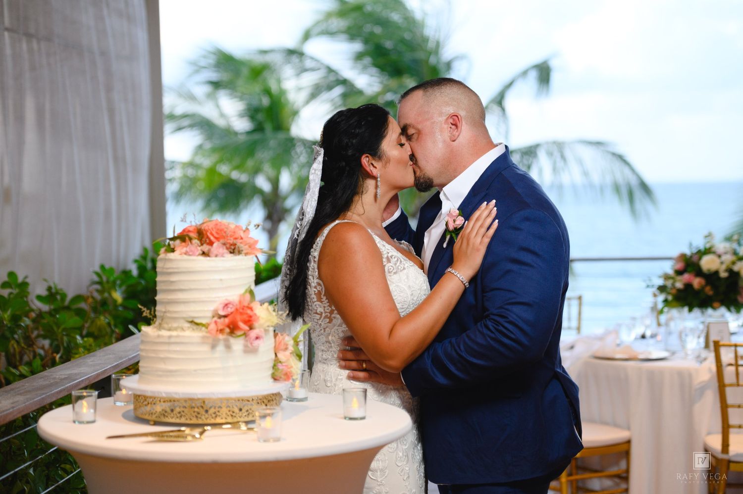 Bride and groom share a kiss beside their elegant white wedding cake with pink flowers at an oceanfront venue.