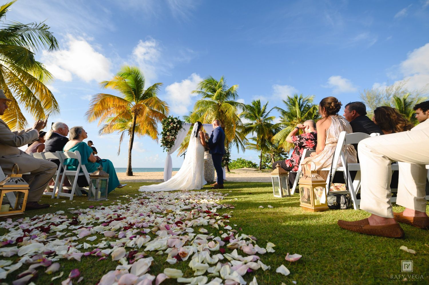 Outdoor beach wedding ceremony with rose petal aisle, palm trees, and guests seated on white chairs under a sunny tropical sky.