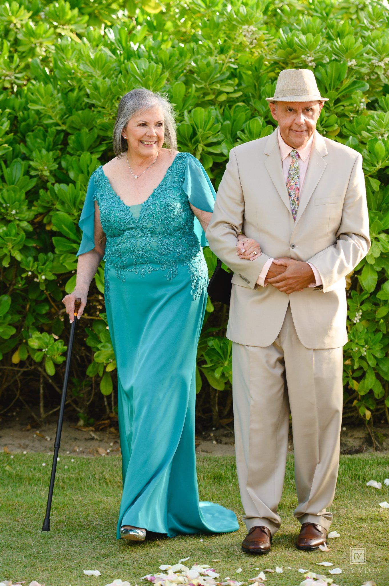 Elderly couple walking outdoors, woman in teal gown with cane, man in beige suit with floral tie.