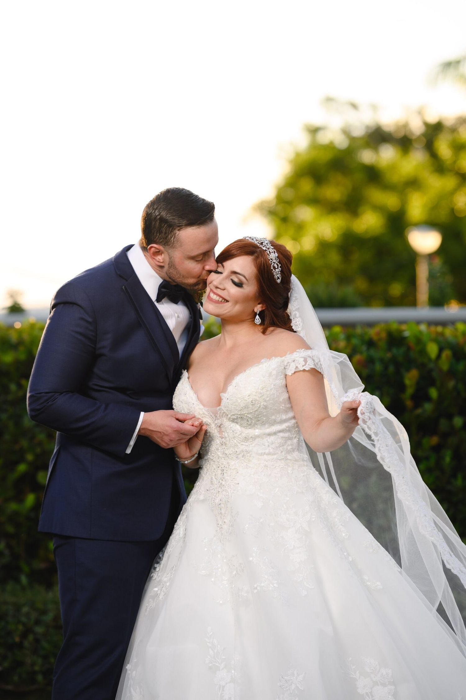 Groom in navy tuxedo kissing bride in lace off-shoulder wedding gown at golden hour outdoors.