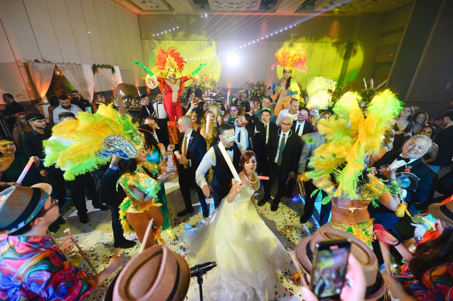 Bride and groom surrounded by colorful carnival dancers with feathered costumes at a festive wedding reception.