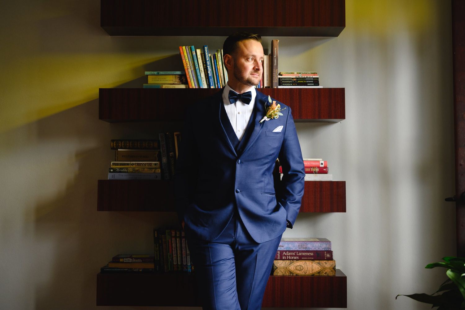 Groom in navy blue three-piece suit with bow tie and boutonniere poses confidently in front of wooden bookshelves.