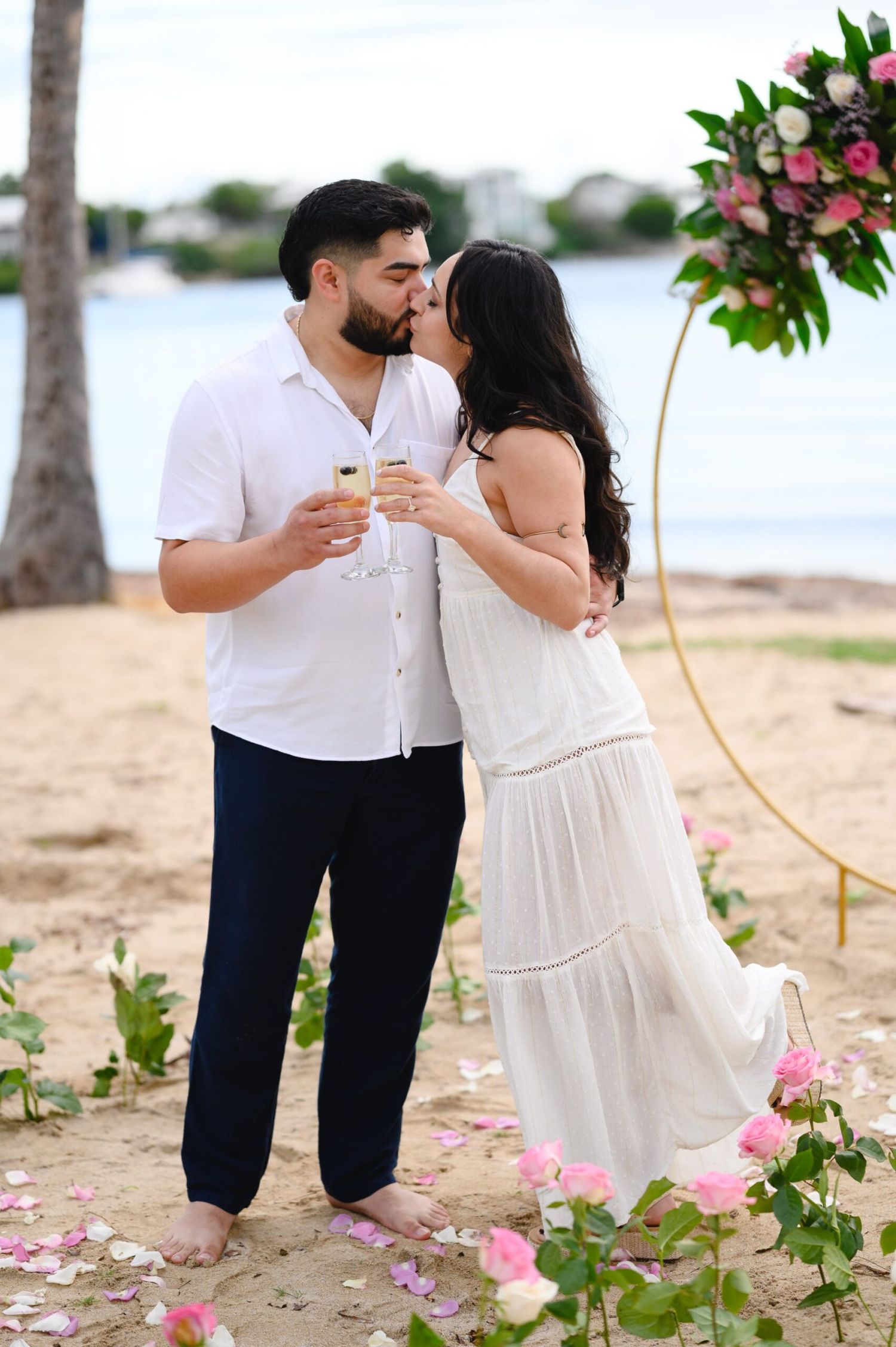 Couple shares romantic kiss under circular floral arch on sandy beach with rose petals.